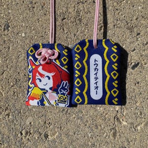 May include: Two rectangular fabric omamori charms with pink hanging cords. One features an embroidered red-haired anime character in a blue uniform, while the other displays a white vertical label with the Japanese text トウカイテイオー.