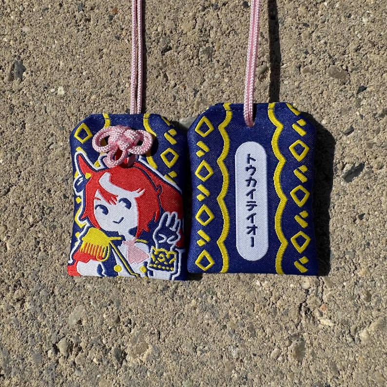 May include: Two rectangular fabric omamori charms with pink hanging cords. One features an embroidered red-haired anime character in a blue uniform, while the other displays a white vertical label with the Japanese text トウカイテイオー.