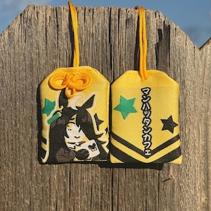 May include: Two yellow fabric omamori charms with yellow hanging cords. One charm features an embroidered anime-style character with black hair and a white book. The other charm displays green and black stars and Japanese text reading マンハッタンカフェ.