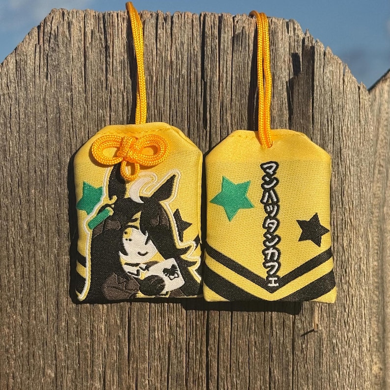 May include: Two yellow fabric omamori charms with yellow hanging cords. One charm features an embroidered anime-style character with black hair and a white book. The other charm displays green and black stars and Japanese text reading マンハッタンカフェ.