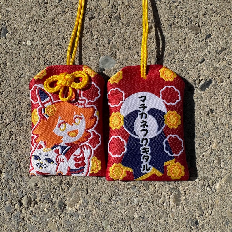 May include: Two rectangular red fabric omamori charms with yellow hanging cords. The left charm features an embroidered orange-haired character and a white cat. The right charm displays a white circular emblem with Japanese text.