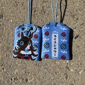 May include: Two light blue fabric omamori charms with light blue hanging cords. One charm features an embroidered anime-style character with brown hair and a rose, while the other displays red and blue rose embroidery and the Japanese text Rice Shower.
