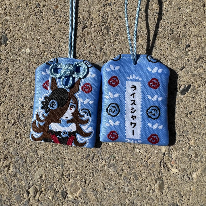 May include: Two light blue fabric omamori charms with light blue hanging cords. One charm features an embroidered anime-style character with brown hair and a rose, while the other displays red and blue rose embroidery and the Japanese text Rice Shower.