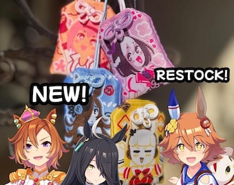 May include: Four rectangular fabric omamori charms featuring embroidered anime-style character illustrations in orange, purple, blue, and yellow. The charms have decorative knotted cords at the top. Text overlays read NEW! and RESTOCK!