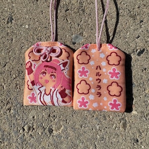 May include: Two peach-colored fabric omamori charms with pink cords. One charm features an embroidered anime-style character with pink hair, while the other displays pink flower motifs and the Japanese text "ハルウララ" in dark brown.