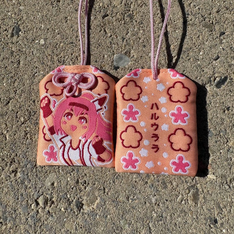 May include: Two peach-colored fabric omamori charms with pink cords. One charm features an embroidered anime-style character with pink hair, while the other displays pink flower motifs and the Japanese text "ハルウララ" in dark brown.