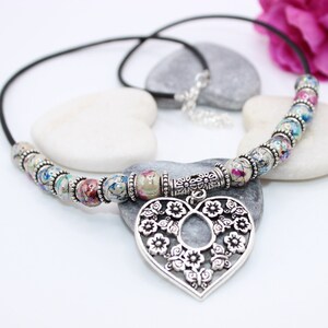 May include: A black leather necklace with a silver heart pendant. The pendant is decorated with floral designs and the necklace is adorned with colorful beads.