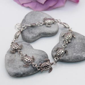 May include: Silver-toned anklet featuring a series of detailed turtle charms. The charms are connected by delicate links, creating a charming and whimsical accessory. The anklet is displayed on a light background with gray stones and a purple flower.