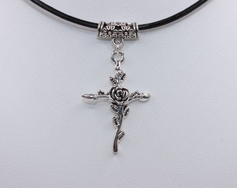 May include: A silver-toned metal cross pendant featuring a central rose and leaf design, suspended from a black cord by a cylindrical bail decorated with a small flower pattern. The cross has a textured finish and is centered on a plain white surface.