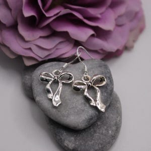 May include: Silver-toned bow-shaped dangle earrings. The earrings are displayed on gray stones, with a blurred purple flower in the background. The earrings have a detailed bow design with a hook closure.