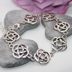 Könnte beinhalten: Ein Armband aus silberfarbenem Metall mit einer Reihe von sich wiederholenden, durchbrochenen keltischen Knotenanhängern, die durch kleine kreisförmige Verbindungsringe verbunden sind. Das Armband hat einen Karabinerverschluss.