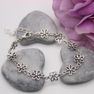 Pode incluir: Uma pulseira prateada com um design de flores de margarida. A pulseira é composta por vários berloques em forma de margarida ligados entre si. A pulseira é exibida em pedras cinzentas com uma flor roxa ao fundo.