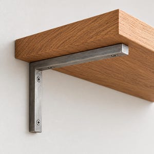 Puede incluir: Un estante de madera de color marrón claro con un patrón de veta natural prominente, sostenido por un soporte de metal cepillado en forma de L. El soporte está fijado firmemente a la parte inferior del estante y a la pared con tornillos.