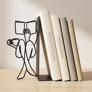 Può includere: Un fermalibri in metallo nero con la silhouette a linea continua di una persona seduta che legge un libro e tiene in mano una tazza. Il fermalibri sostiene quattro libri con i titoli Selected Essays e Philosophy.