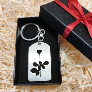 Può includere: Un portachiavi in ​​argento con un design a forma di rosa, presentato in una scatola regalo nera con un nastro rosso.