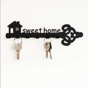 May include: Black metal key holder shaped like a key, with the words "sweet home" and a house silhouette. Includes six key hooks and two sets of keys hanging from the hooks. The key holder is mounted on a white wall.