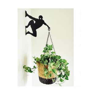 May include: Black metal wall-mounted plant hanger shaped like a superhero figure holding a chain with a hanging plant pot. The pot is made of natural fiber and has a black base. The plant is a green leafy vine.
