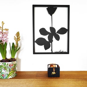 May include: A black framed wall art piece featuring a silhouette of a rose with the text "voulez-vous" at the bottom. A potted plant with pink flowers and a small black metal candle holder are on a wooden shelf below the art.