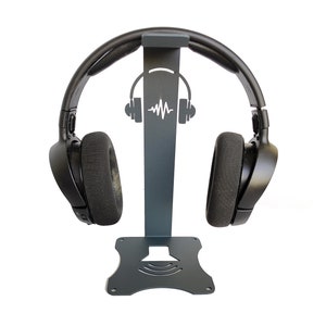 Op de afbeelding: Een zwarte metalen hoofdtelefoonstandaard met een geluidsgolfontwerp. De standaard heeft een ronde basis en een verticale paal met een haak aan de bovenkant om de hoofdtelefoon vast te houden.