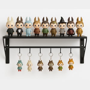Puede incluir: Una estantería de metal negro exhibe diez figuras coleccionables con orejas de conejo y llaveros a juego. Las figuras visten varios atuendos, incluyendo camisas a rayas, una calavera y un sombrero de bruja. Los llaveros reflejan los diseños de las figuras.