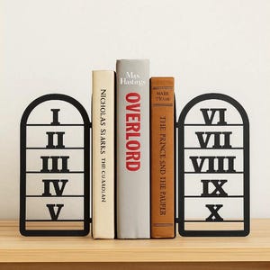 May include: Black metal bookends with Roman numerals I-V and VI-X, holding three books. The books are titled "Overlord", "The Prince and the Pauper", and "Nicholas Sparks The Guardian". The bookends are on a wooden surface.