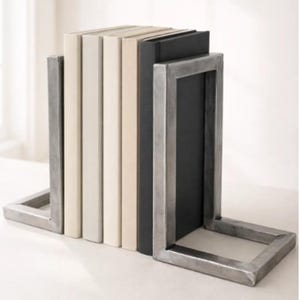Puede incluir: Un par de sujetalibros de metal en forma de L con acabado en plata cepillada, que sostienen una fila de cinco libros. Los libros tienen cubiertas sólidas en tonos crema, beige y negro, dispuestos verticalmente entre los soportes metálicos.