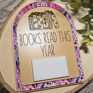 May include: Wooden plaque with the text "BOOKS READ THIS YEAR" and a row of illustrated books. The plaque has a decorative border with a colourful pattern and a white rectangle for notes. The plaque is on a wooden surface.