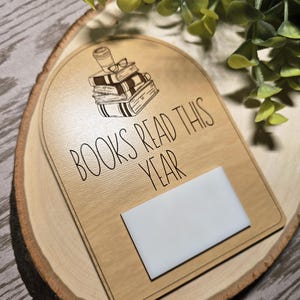 May include: Wooden plaque with the text "BOOKS READ THIS YEAR" and an illustration of books, glasses, and a coffee cup. The plaque has a white rectangle for notes and is set on a wooden slice.