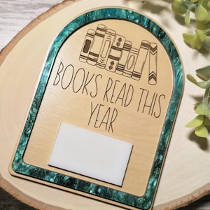 Può includere: Targa in legno con bordo in resina turchese e la scritta "BOOKS READ THIS YEAR". La targa presenta un'illustrazione di libri impilati e un rettangolo bianco vuoto per le note. La targa è posta su una fetta di legno.