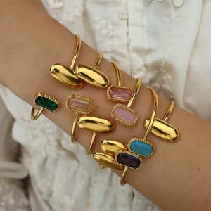 May include: Gold-coloured bracelets and rings with colourful rectangular gemstones. The bracelets have a curved design with a large, smooth, gold-coloured oval at the end. The rings feature gemstones in various colours, including green, pink, and blue.