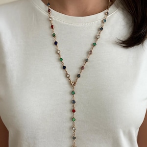 May include: A gold-coloured necklace with a Y-shaped pendant. The necklace features a series of colourful, round gemstones in red, blue, green, and clear settings. The necklace is worn against a white t-shirt.
