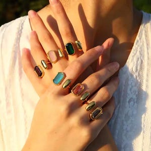 May include: Gold-plated rings with rectangular gemstones in various colours. The rings feature gemstones in pink, green, blue, and purple. The gemstones are set in a gold band, creating a stylish look. The rings are displayed on a person's fingers.