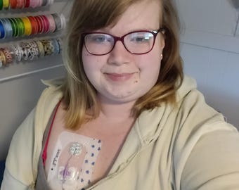 May include: A beige zip-up hoodie featuring a wide, horizontal chest opening designed for medical access. The garment is worn over a gray tank top, revealing a clear adhesive medical dressing with a central port and purple connector tubing.