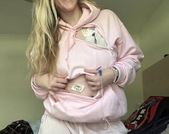 May include: A light pink hooded sweatshirt featuring a zippered opening on the chest area to accommodate medical tubing and a central abdominal access point. The garment includes a drawstring hood, long sleeves, and a front kangaroo pocket.