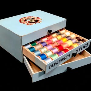 May include: A light blue wooden storage cabinet with two drawers containing rows of colorful sewing thread spools. The top drawer is open, revealing a grid of thread. The text "DEWHURST'S SYLKO" is printed on the drawer front, with a circular logo on top.