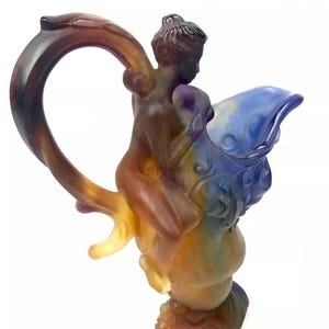 May include: A glass pitcher with a blue and brown iridescent finish. A nude figure is riding on the pitcher's handle, which is shaped like a branch.