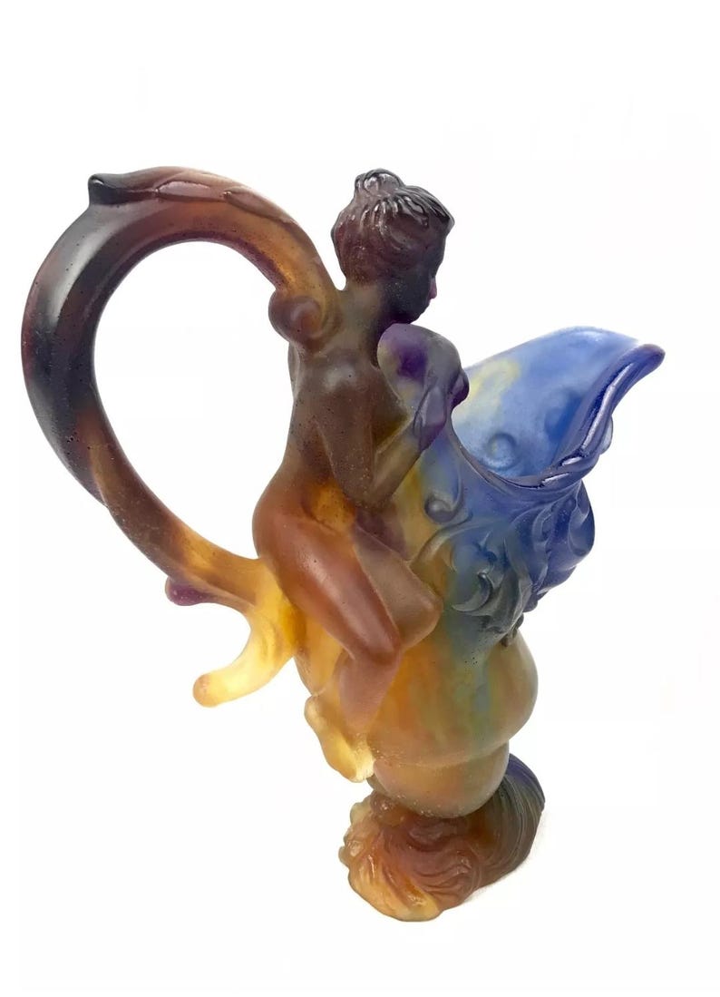 May include: A glass pitcher with a blue and brown iridescent finish. A nude figure is riding on the pitcher's handle, which is shaped like a branch.