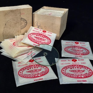 May include: A stack of white paper squares with red text that reads "Transparent 87180 Progress Made in England Unbreakable Squares 25/1000" and a clear glass circle.