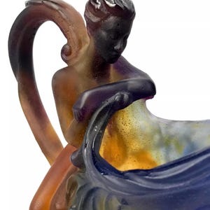 May include: A glass pitcher with a handle shaped like a woman holding a large shell. The pitcher is made of iridescent glass with shades of purple, blue, yellow, and brown.