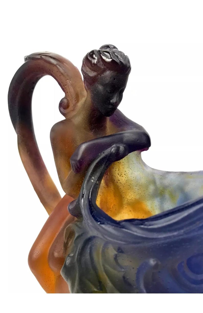 May include: A glass pitcher with a handle shaped like a woman holding a large shell. The pitcher is made of iridescent glass with shades of purple, blue, yellow, and brown.