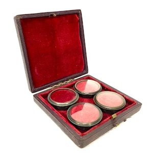 Könnte beinhalten: Eine antike Holzkiste mit rotem Samt-Innenfutter, die vier kleine Glasbehälter mit roten Deckeln enthält. Die Kiste ist geöffnet und die Behälter sind sichtbar.