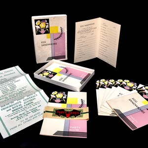 May include: A vintage game box set called "Egg Sandwiches" with a pink and black design. The box contains cards with floral illustrations and a booklet with instructions. The game is a party game.
