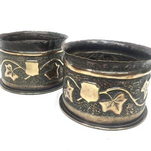 May include: Two antique metal napkin rings. The rings are cylindrical with a dark, textured exterior and a gold-coloured band. The design includes gold-coloured leaves and shield-shaped accents. The rims have a decorative, uneven edge.