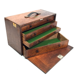 May include: A vintage wooden tool chest with a black handle and three open drawers. The drawers have green felt lining and black knobs. The chest is made of dark brown wood and has a hinged bottom.