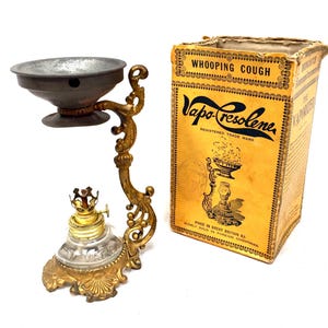 Può includere: Un vaporizzatore Vapo-Cresolene vintage con supporto in metallo dorato, base in vetro trasparente e ciotola in metallo scuro. Accanto si trova la scatola di cartone gialla originale con testo nero che recita Whooping Cough e Vapo-Cresolene.