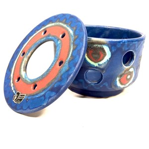 May include: Blue and red ceramic tea warmer with a lid. The warmer has a circular design with holes for heat circulation. The lid has a ring with a smaller hole in the centre.