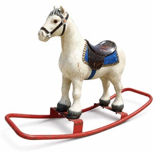 May include: A vintage rocking horse with a white body, dark grey hooves, and a brown bridle. It features a dark brown saddle with blue trim and is mounted on a red metal frame. The paint shows signs of wear and age throughout the entire structure.