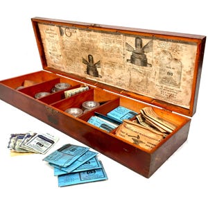 Può includere: Una scatola di legno piena di piccoli pacchetti di cristalli per orologi. La scatola è etichettata "Genuine G.S. Dichroic Crystals" e "Wearable Watch Crystals First in 1928."