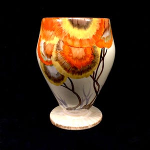 May include: A decorative ceramic vase with a cream-colored base and body. The vase features a painted design of orange, yellow, and brown floral patterns. Brown branches extend from the base, adding to the artistic appeal. The vase stands on a round, cream-colored base.
