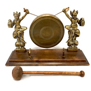 May include: A brass gong set featuring two figures perched on mushrooms, each holding a striker toward a central circular gong. The figures are mounted on a rectangular wooden base with two small metal hooks. A separate wooden mallet with a rounded head is included.
